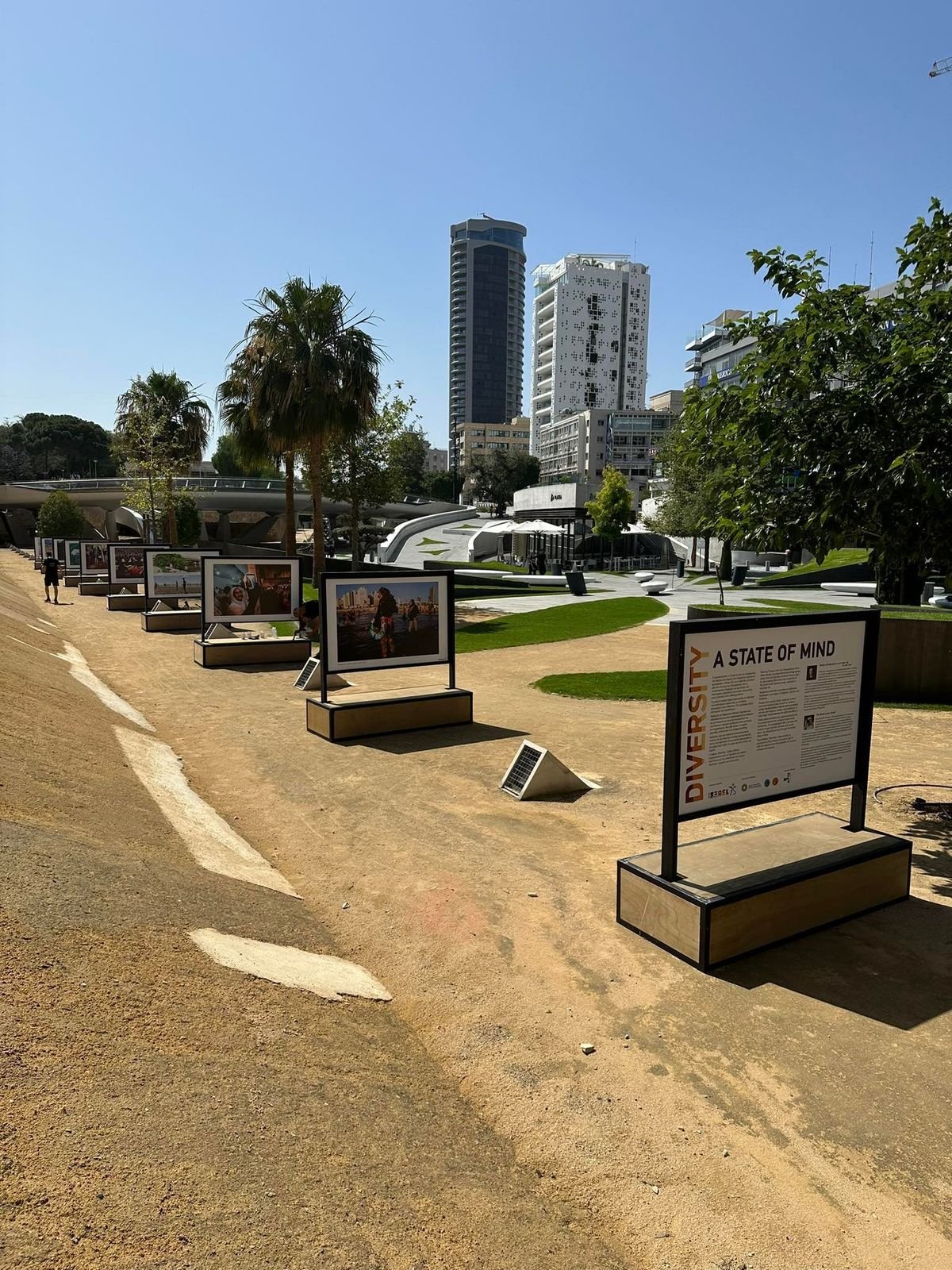 Row of outdoor exhibition display stands set up in a park in central Nicosia, with city towers behind